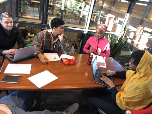 Four people sitting at a table and having a discussion. Two of them are typing on a laptop.