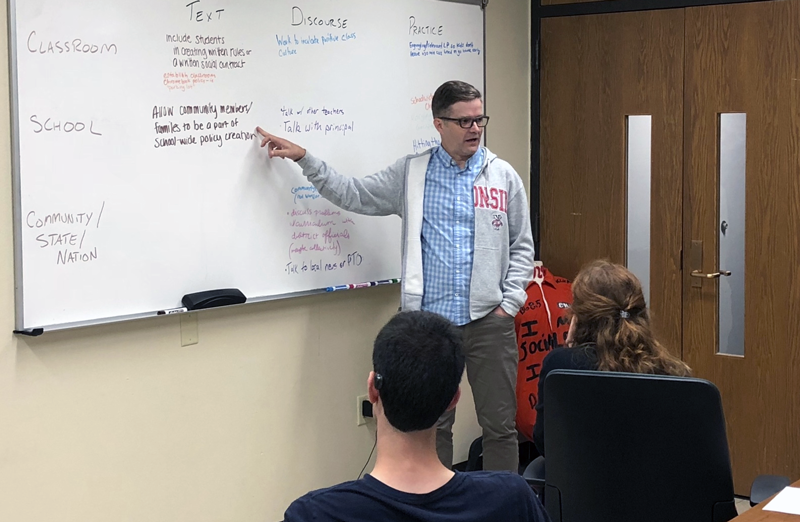 Teacher in classroom pointing at text on a whiteboard while looking at the class