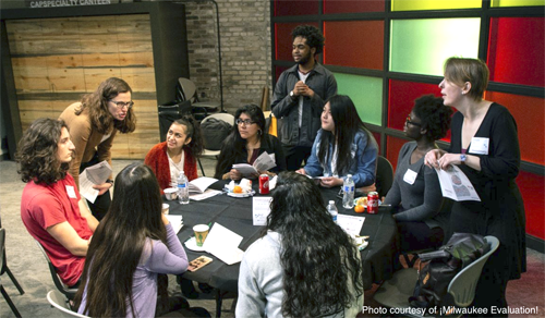 A group of people sitting around a table. One person is standing and leaning over while talking to the group.